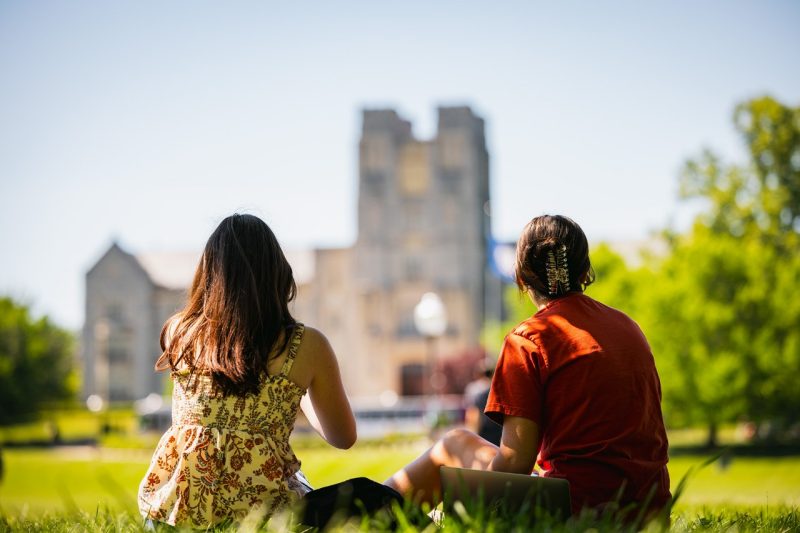 Two students sit on the lawn at Virginia Tech looking towards a blurred image of Burress Hall